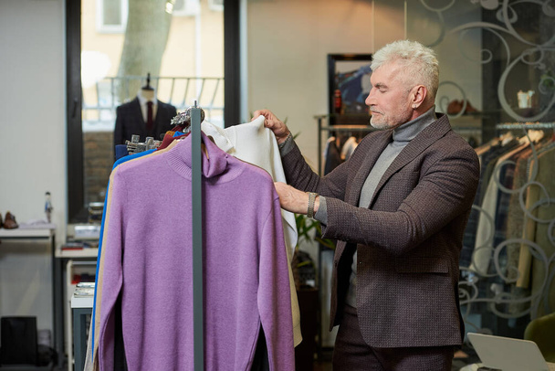 Ένας ώριμος άντρας με γκρίζα μαλλιά διαλέγει πουλόβερ ζιβάγκο σε κατάστημα ρούχων. Ένας άντρας πελάτης με μάλλινο κοστούμι σε μπουτίκ.. - Φωτογραφία, εικόνα