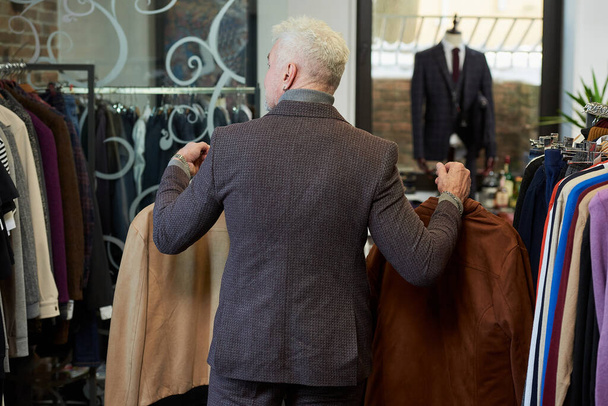 Egy ősz hajú és sportos fizikumú felnőtt férfi fényképe megy két kabáttal a ruhaüzlet próbafülkéjébe. Egy szakállas férfi öltönyt visel egy butikban. - Fotó, kép