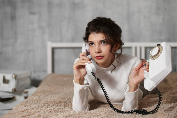 Nuori nainen makaa sängyssä ja puhuu puhelimessa kotona, mietteliäs nuori tyttö pitelee puhelinta, kaunis teini pitää vintage-puhelinta kädessään.. - Valokuva, kuva