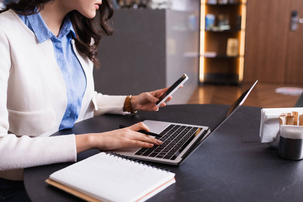 visão parcial do freelancer segurando smartphone enquanto digita no laptop no café - Foto, Imagem