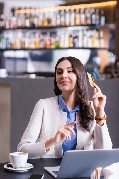 freelancer sonriente mirando hacia otro lado y señalando con el dedo mientras sostiene la tarjeta de crédito en el restaurante - Foto, imagen
