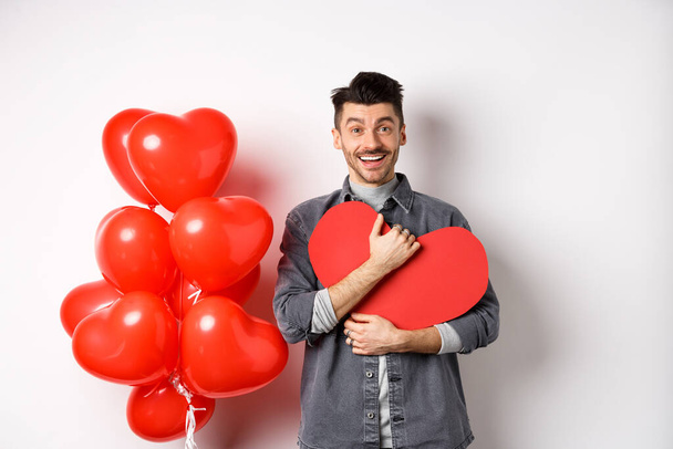 Young romantic man looking with love at camera, hugging Valentine heart card and smiling happy, celebrating lovers holiday, standing near red balloons, white background - Fotó, kép
