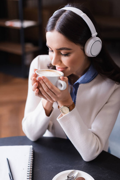 mulher satisfeita em fones de ouvido sem fio desfrutando de sabor de café enquanto segurando copo com olhos fechados - Foto, Imagem