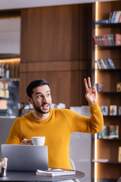 freelancer feliz árabe mostrando gesto de vitória enquanto segurando xícara de café no restaurante - Foto, Imagem