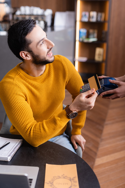 vrolijk Arabisch freelancer houden credit card in de buurt van betaalterminal in handen van serveerster, wazig voorgrond - Foto, afbeelding