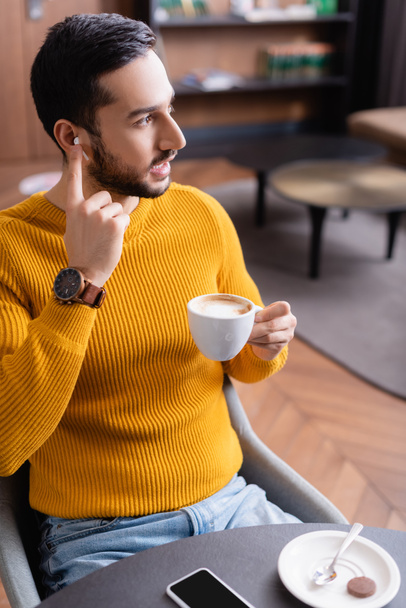 giovane uomo arabo distogliendo lo sguardo mentre regola auricolare e tenendo in mano una tazza di caffè nel ristorante - Foto, immagini