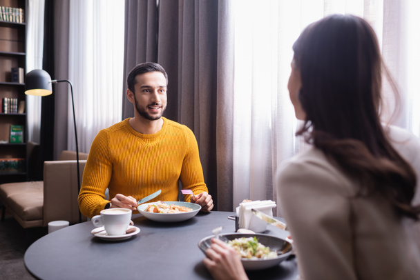Hombre árabe sonriendo a su novia en primer plano borroso cerca de la comida y el café en el restaurante  - Foto, Imagen