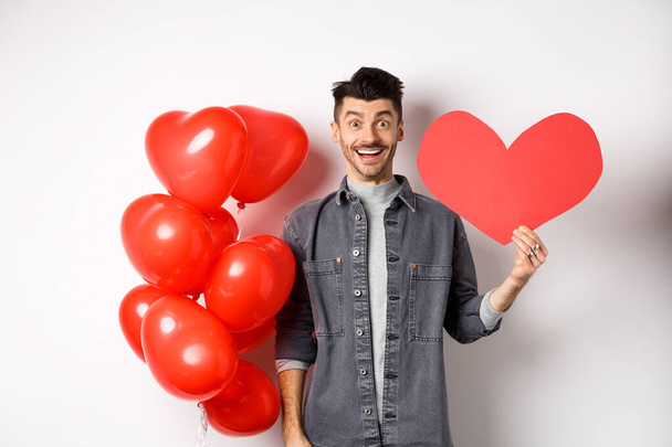 Saint Valentin et concept d'amour. Joyeux gars drôle montrant découpe de coeur, debout près de ballons romantiques et souriant excité à la caméra, fond blanc - Photo, image