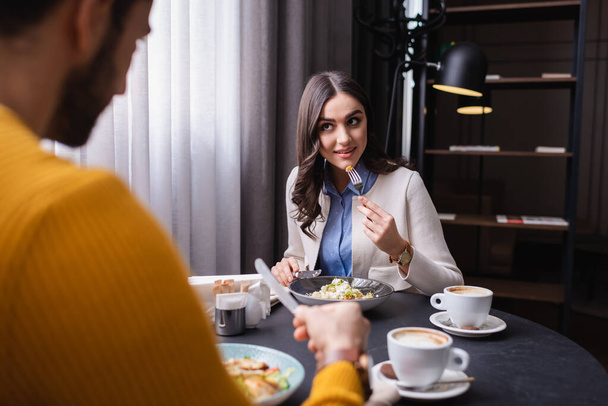Jovem mulher olhando para o namorado em primeiro plano borrado perto de salada saborosa e café no restaurante  - Foto, Imagem