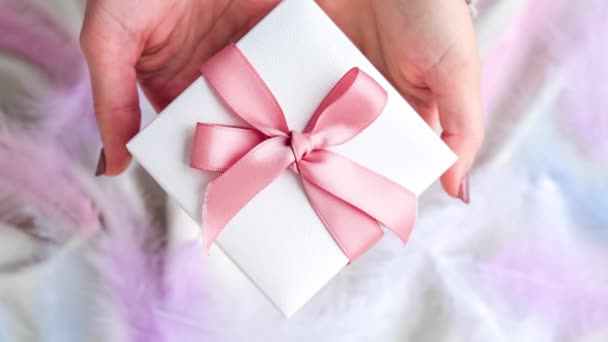 Weibliche Hand hält weiße Geschenkschachtel mit rosa Schleife auf weißem Seidenhintergrund. Bunte Vogelfedern. Ferienkonzept. 4k Video. Vergrößern - Filmmaterial, Video