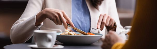 Καλλιεργημένη άποψη της γυναίκας κρατώντας μαχαιροπήρουνα κοντά στη σαλάτα, κύπελλο και το φίλο σε θολή πρώτο πλάνο στο εστιατόριο, πανό  - Φωτογραφία, εικόνα