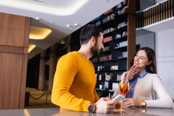 Веселая женщина, стоящая рядом с арабским другом со смартфоном и напитками на барной стойке  - Фото, изображение