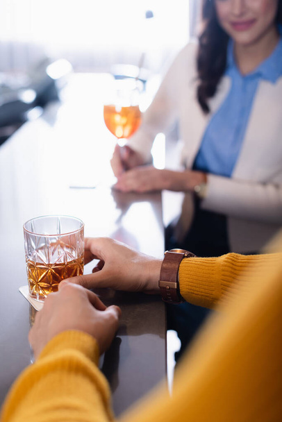 Vista recortada del vaso de whisky en la mano del hombre cerca de amigo sobre fondo borroso en el restaurante  - Foto, Imagen