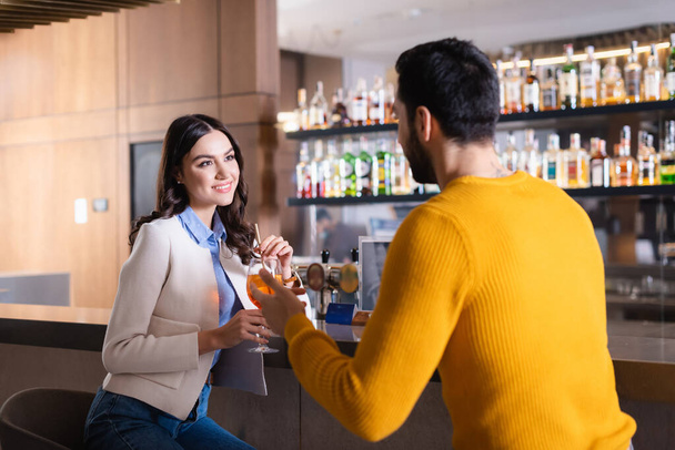 glimlachende vrouw met cocktail kijken naar moslim vriend op wazig voorgrond in de buurt bar teller in restaurant  - Foto, afbeelding