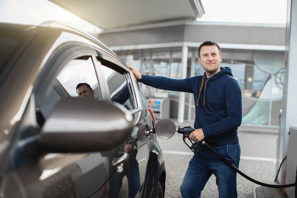 Young cheerful Caucasian man in casual outfit, refueling his modern luxury car at fuel station, smiling to camera - Foto, afbeelding