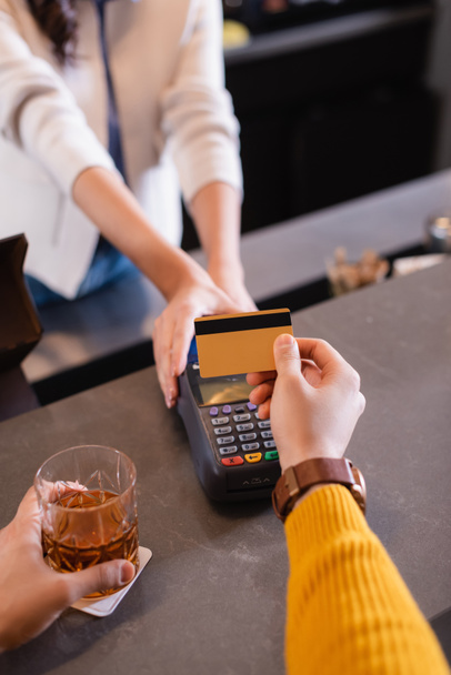 Cropped view of man with whiskey paying with credit card near bartender with terminal on blurred background  - Photo, Image