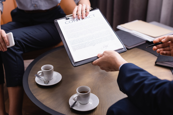Cropped view of businessman holding contract near businesswoman and coffee on blurred background  - Photo, Image
