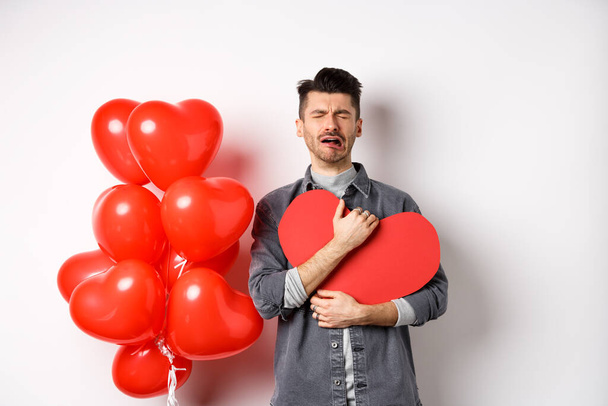 Crying man standing single and lonely on Valentines day, hugging heart cutout and sobbing miserable, being heartbroken and rejected by lover, white background - Fotó, kép