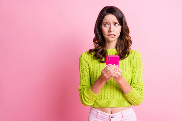 Foto van nerveus bang meisje houden mobiele telefoon beet lip look lege ruimte dragen groene trui geïsoleerde roze kleur achtergrond - Foto, afbeelding