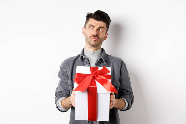 Θλιμμένος φίλος κοιτάζοντας στην άκρη και κρατώντας κουτί δώρου, περιμένοντας για τον εραστή, κάνοντας δώρο έκπληξη την ημέρα του Αγίου Βαλεντίνου, ημερομηνία σχεδιασμού με τη φίλη, λευκό φόντο - Φωτογραφία, εικόνα