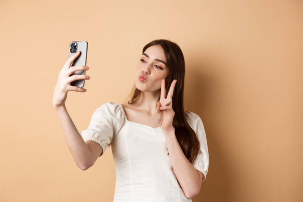 ファッション女性ブロガーパッカーの唇やスマートフォンのカメラでv-signを表示,ソーシャルメディアのために自画自身を取ります,ベージュの背景に立って - 写真・画像