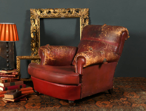 Old shabby κόκκινη δερμάτινη πολυθρόνα τοποθετημένη κοντά σε vintage διακοσμητικά πλαίσια και λάμπα στο δωμάτιο με ρετρό στυλ εσωτερικό - Φωτογραφία, εικόνα