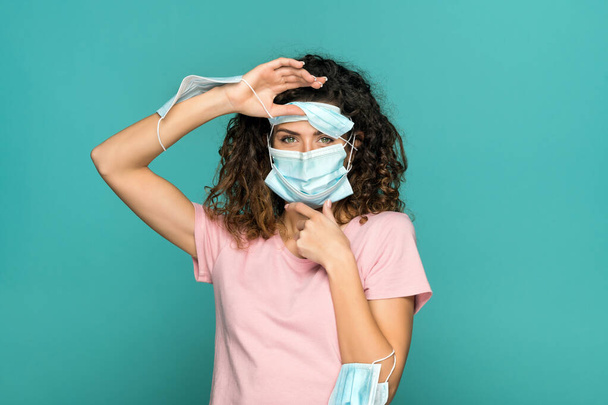 Mujer bonita con muchas máscaras médicas en la cara y los brazos mientras protege contra el coronavirus y mira a la cámara en el fondo azul en el estudio - Foto, imagen