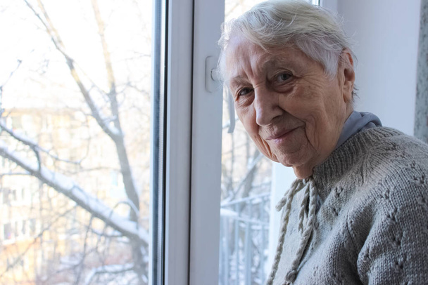 Yaşlı yalnız kadın evinin penceresinin yanında oturuyor ve kışın kameraya bakıyor ve gülümsüyor. - Fotoğraf, Görsel