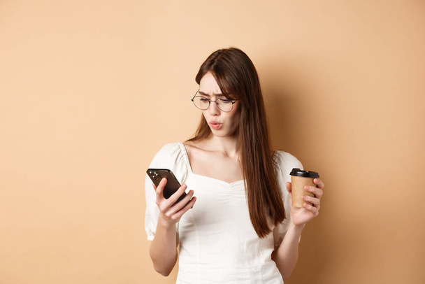 Mujer joven conmocionada leyendo la pantalla del teléfono móvil y frunciendo el ceño, sosteniendo la taza de café, de pie sobre un fondo beige - Foto, imagen