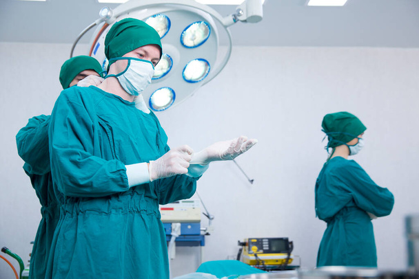 青い外科用ガウンの外科医は、緊急時のためのマウスガードを結合します。外科医は現代医療機器を備えた手術室で手袋を着用しています. - 写真・画像