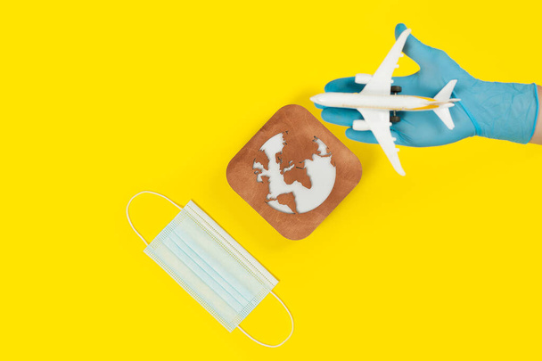 Plane model in hand, face mask  and earth model on a yellow background. Hands in gloves. Flight impact of coronavirus (COVID-19) concept. Afric - Фото, изображение