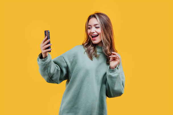 手に携帯電話を持って黄色の背景に魅力的な女の子、彼女は感情的に電話と笑顔を見ています。広告用の写真。スタジオ写真. - 写真・画像