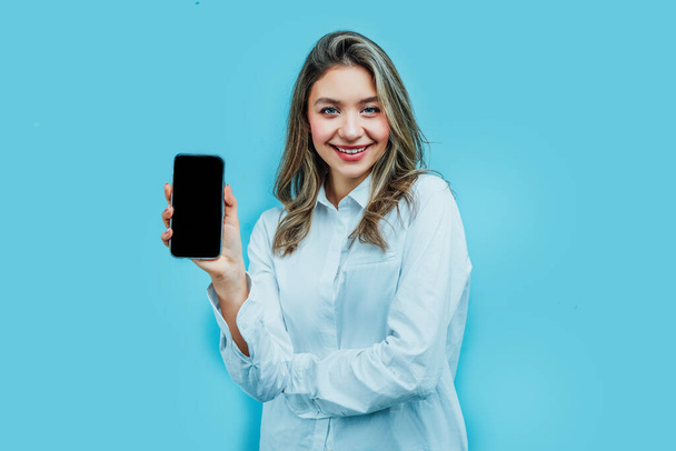Portrait of an attractive girl holding a phone with a black screen in her hand, behind her a blue background. Studio photo. Commerce. - Photo, Image