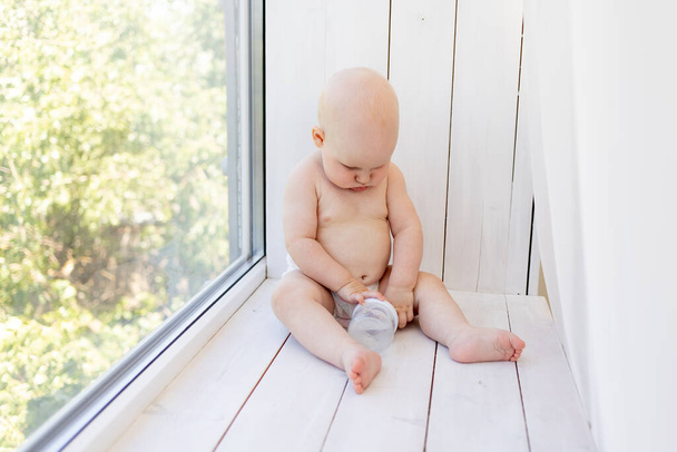 baby on the window with a bottle of milk in diapers at home - 写真・画像