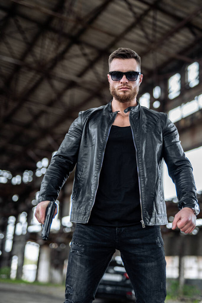 Handsome young man in leather jacket holding a gun. Cool sunglasses on brutal man. Brutal male in abandoned warehouse. - 写真・画像