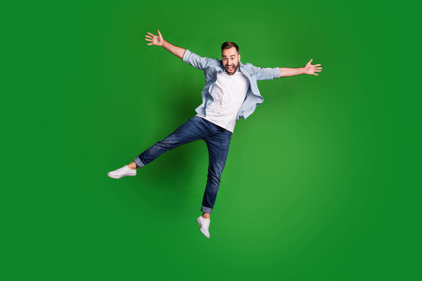 Full size photo of optimistic funky guy jump yell wear blue shirt jeans footwear isolated on green color background - Zdjęcie, obraz