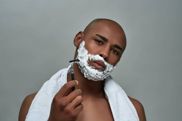 Handsome young african american man with shaving foam applied on his face looking at camera, holding steel razor, posing isolated over gray background - Foto, Imagem