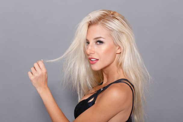 Beauty profile portrait. Hot blonde girl in black bra, on grey background, arranges her hair, looking at camera. - 写真・画像