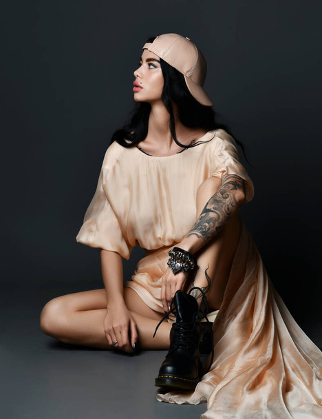 Young sexy brunette woman with tattoo on arm and piercing on face in dress and massive boots sitting on floor and looking aside - Foto, afbeelding