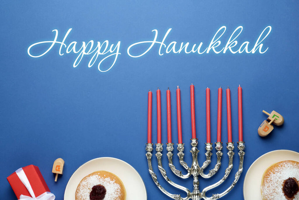 Happy Hanukkah. Traditional menorah, candles, sufganiyot and dreidels on blue background, flat lay  - Photo, image
