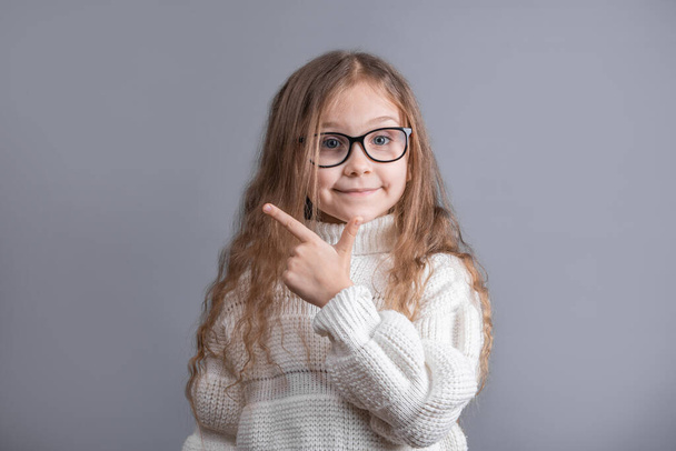 Портрет молодой привлекательной маленькой девочки с длинными светлыми волосами в белом свитере улыбается и указывает в сторону на сером фоне студии. Место для смс. Копирование пространства - Фото, изображение