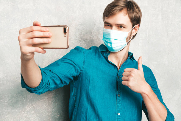 Handsome smiling hipster lumbersexual businessman model wearing casual jeans shirt clothes. Fashion stylish man posing against gray wall in studio. In protective mask for corona virus. Taking selfie  - Foto, Imagen