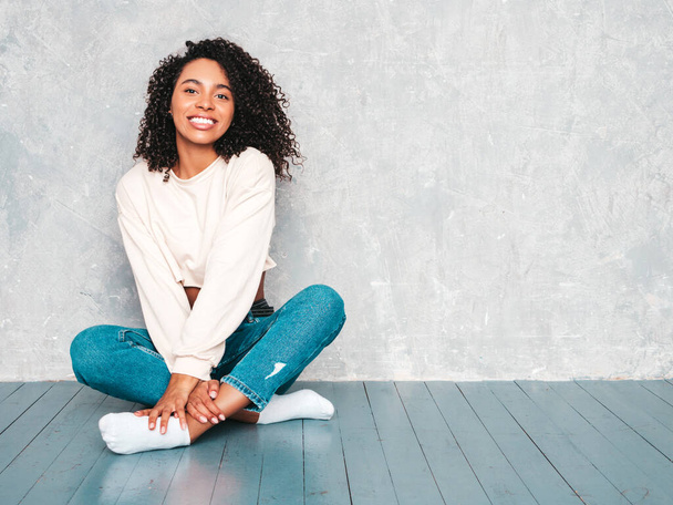 Beautiful black woman with afro curls hairstyle.Smiling model in trendy jeans clothes and sunglasses. Sexy carefree female sitting on the floor near gray wall in studio - Фото, изображение