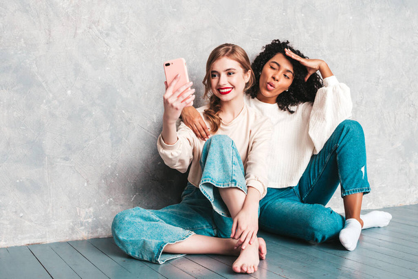 Two young beautiful smiling international hipster female in trendy summer jeans clothes. Sexy carefree women sitting near gray wall in studio. Positive models having fun. They taking selfie photos - 写真・画像