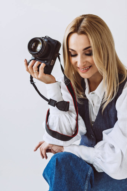 A beautiful woman photographer in a denim casual outfit and a white blouse with voluminous sleeves with a camera in her hands. Hobbies. Soft selective focus. - Zdjęcie, obraz