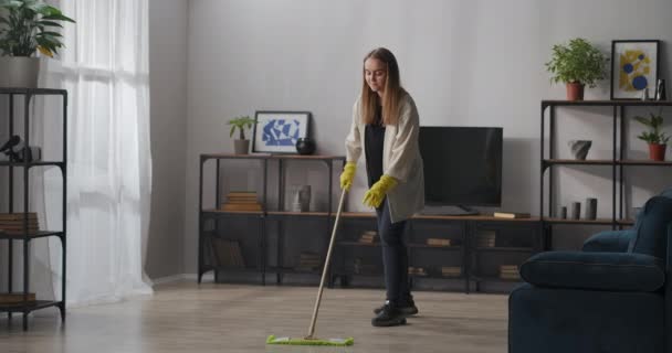 mujer rubia joven está lavando el piso en el apartamento en el día soleado, usando fregona y guantes de goma, servicio de limpieza - Metraje, vídeo