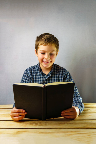 Uma criança feliz lê um livro. Um menino de 7-9 anos de idade em uma camisa xadrez em uma mesa de madeira com um livro. Dia Mundial do Livro - Foto, Imagem