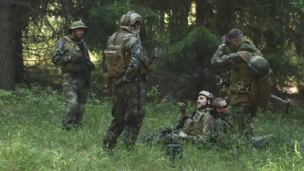 Полный снимок взвода вооруженных солдат в камуфляжной форме, отдыхающих перед военной операцией, лежащих на траве в лесу, курящих сигареты и беседующих - Кадры, видео