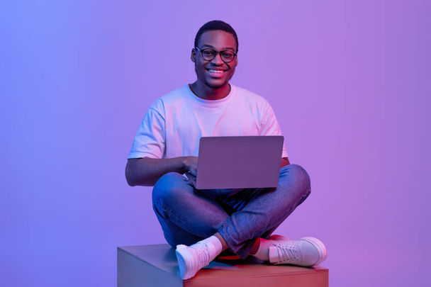 Glücklich Schwarz Student Guy In Brille Sitzen Mit Laptop In Neon Lighting - Foto, Bild