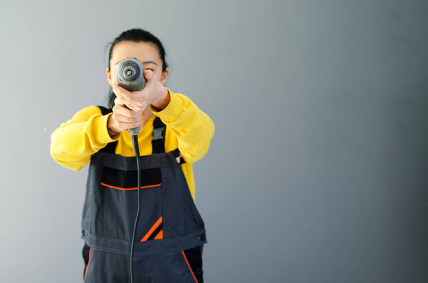 Female construction worker dressed in special clothes with a cordless screwgun, a construction tool isolated on grey background. Asian Korean woman with drill - Zdjęcie, obraz
